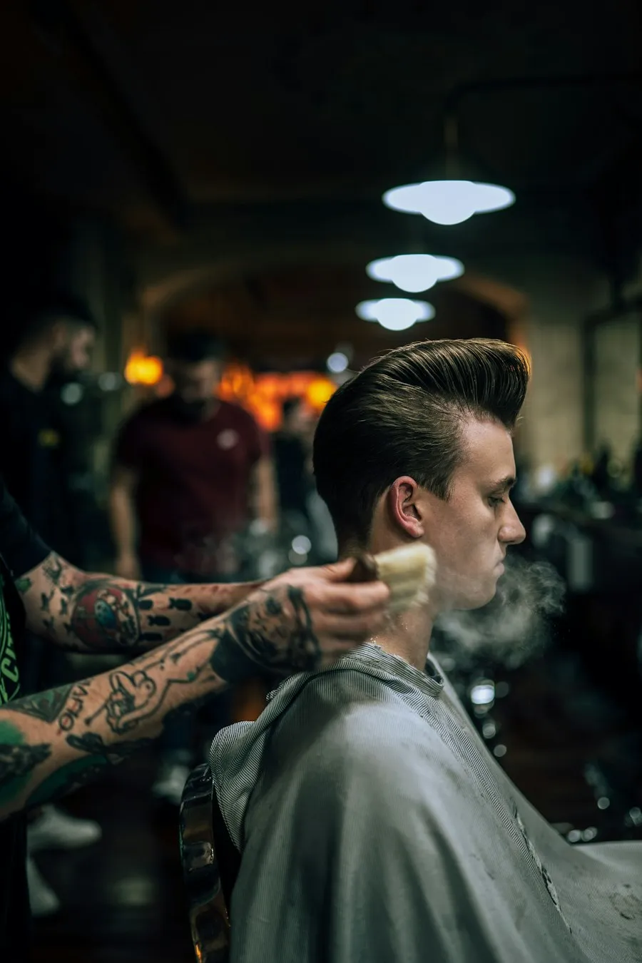 Cliente na cadeira enquanto o barbeiro, com o braço tatuado, finaliza o corte