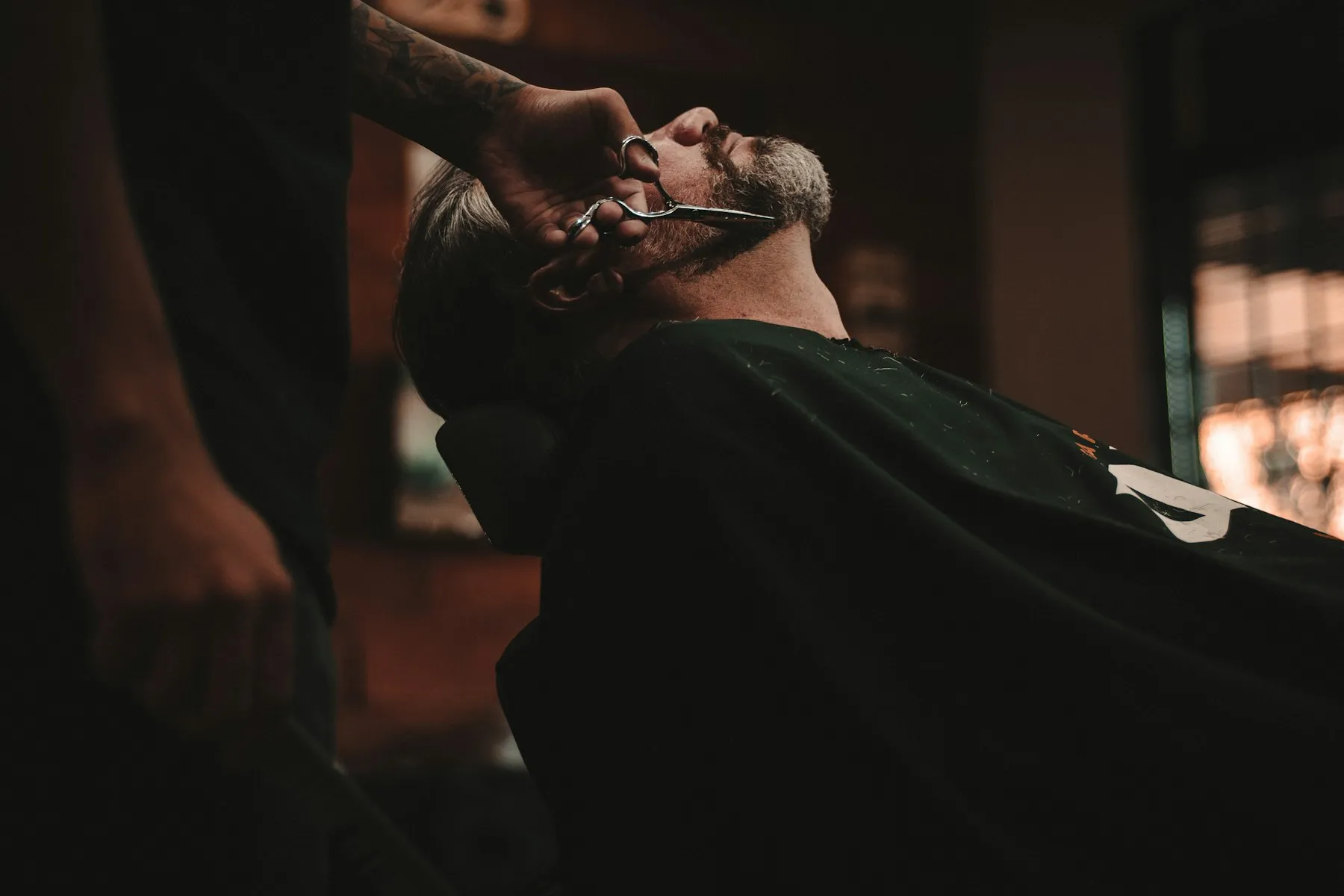 Barbeiro aparando a barba de um cliente reclinado na cadeira, em ambiente de luz baixa